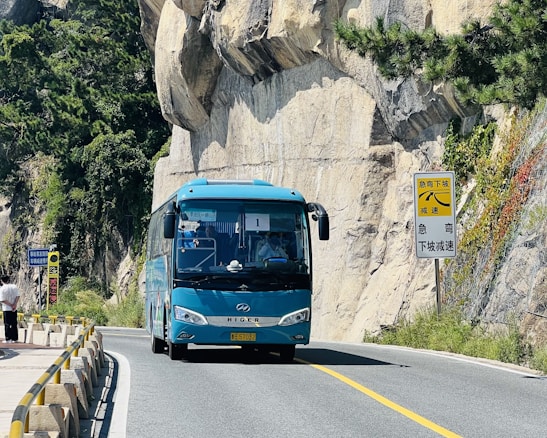 A blue tour bus is traveling on a winding mountain road with rocky cliffs on one side. Tall green trees and shrubs are visible alongside the road. There are road signs indicating a sharp turn ahead.