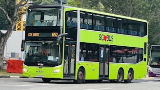A bright green double-decker bus with black accents is driving on a city street. The bus displays the route 'City Direct 652' and features the SG Bus logo with a red heart symbol. There are trees and construction equipment in the background.