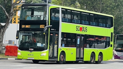 A bright green double-decker bus with black accents is driving on a city street. The bus displays the route 'City Direct 652' and features the SG Bus logo with a red heart symbol. There are trees and construction equipment in the background.