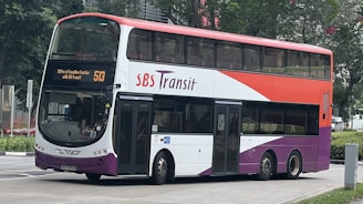 A double-decker bus with a red, white, and purple color scheme featuring the logo 'SBS Transit' on the side. The destination sign on the front of the bus displays route number 513. The bus is parked on a city street with greenery and buildings visible in the background.