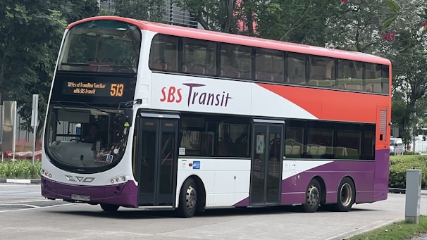 A double-decker bus with a red, white, and purple color scheme featuring the logo 'SBS Transit' on the side. The destination sign on the front of the bus displays route number 513. The bus is parked on a city street with greenery and buildings visible in the background.