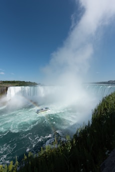 A large waterfall cascades powerfully into a turbulent pool of water below, surrounded by thick mist and a partially visible rainbow. A tour boat, filled with passengers, navigates close to the waterfall, emphasizing the grandeur of the scene. Lush greenery borders the water, contrasting with the bright blue sky above.