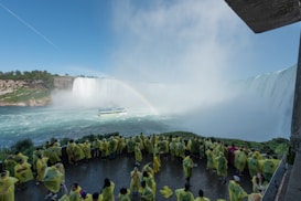 A large group of people wearing yellow rain ponchos stand near a powerful waterfall. A boat sails on the water below the waterfall, and a rainbow is visible in the mist. The surrounding area includes rocky cliffs and lush greenery.