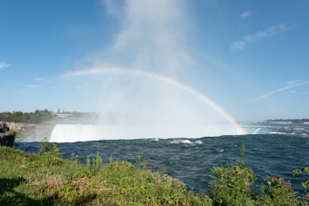 A vibrant rainbow arcs across a misty waterfall with lush greenery in the foreground. The sky is clear and blue, enhancing the brightness of the scene.