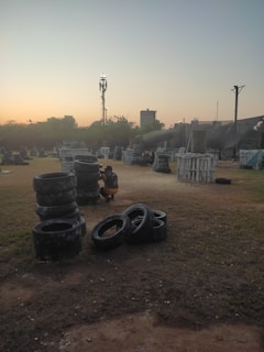 An outdoor paintball field during sunset, featuring stacks of tires and various wooden structures scattered across a grassy area. A person wearing protective gear, including camouflage clothing, is crouched beside a stack of tires. The sky features a gradient of colors from soft orange to blue, indicating early evening or dusk. In the background, there are silhouettes of trees and a few taller structures, including a communication tower.