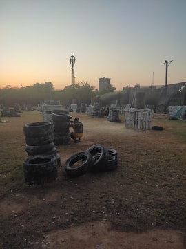 An outdoor paintball field during sunset, featuring stacks of tires and various wooden structures scattered across a grassy area. A person wearing protective gear, including camouflage clothing, is crouched beside a stack of tires. The sky features a gradient of colors from soft orange to blue, indicating early evening or dusk. In the background, there are silhouettes of trees and a few taller structures, including a communication tower.