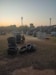 An outdoor paintball field during sunset, featuring stacks of tires and various wooden structures scattered across a grassy area. A person wearing protective gear, including camouflage clothing, is crouched beside a stack of tires. The sky features a gradient of colors from soft orange to blue, indicating early evening or dusk. In the background, there are silhouettes of trees and a few taller structures, including a communication tower.