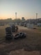 An outdoor paintball field during sunset, featuring stacks of tires and various wooden structures scattered across a grassy area. A person wearing protective gear, including camouflage clothing, is crouched beside a stack of tires. The sky features a gradient of colors from soft orange to blue, indicating early evening or dusk. In the background, there are silhouettes of trees and a few taller structures, including a communication tower.