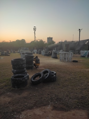 An outdoor paintball field during sunset, featuring stacks of tires and various wooden structures scattered across a grassy area. A person wearing protective gear, including camouflage clothing, is crouched beside a stack of tires. The sky features a gradient of colors from soft orange to blue, indicating early evening or dusk. In the background, there are silhouettes of trees and a few taller structures, including a communication tower.