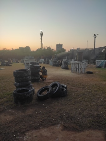 An outdoor paintball field during sunset, featuring stacks of tires and various wooden structures scattered across a grassy area. A person wearing protective gear, including camouflage clothing, is crouched beside a stack of tires. The sky features a gradient of colors from soft orange to blue, indicating early evening or dusk. In the background, there are silhouettes of trees and a few taller structures, including a communication tower.