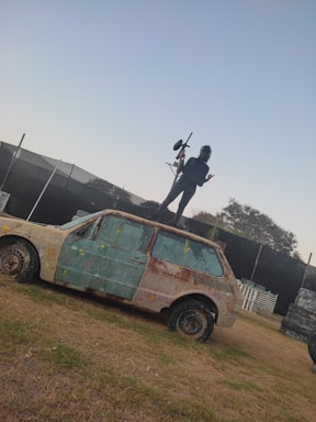 A person wearing protective gear stands on top of an old, rusty car holding a paintball gun. The car is covered in paint splatters and is situated on grassy terrain. In the background, a paintball arena can be seen with barriers and obstacles.