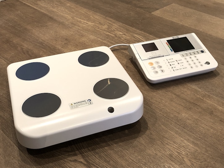 A digital body composition analyzer featuring a square platform with four circular metal sensors and an attached console with a display and various buttons. Both components are white with a sleek, modern design, placed on a wooden floor.