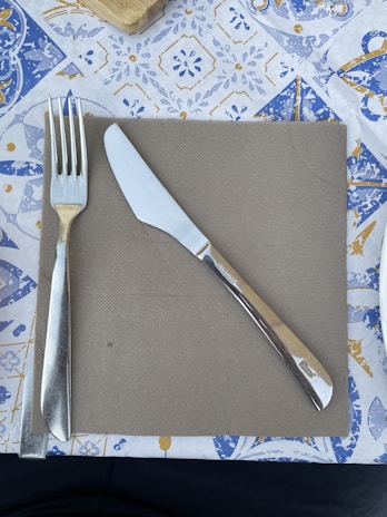 A fork and butter knife are placed on a brown paper napkin, which is on a decorative tablecloth with blue and yellow patterns. The fork is on the left and the knife is on the right, positioned for dining.