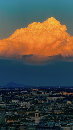 A large, illuminated cloud with vibrant orange hues dominates the sky above a sprawling cityscape. The cloud is detailed with various ridges and textures, while the city below appears mostly in shadow, with buildings and a prominent tower discernible.