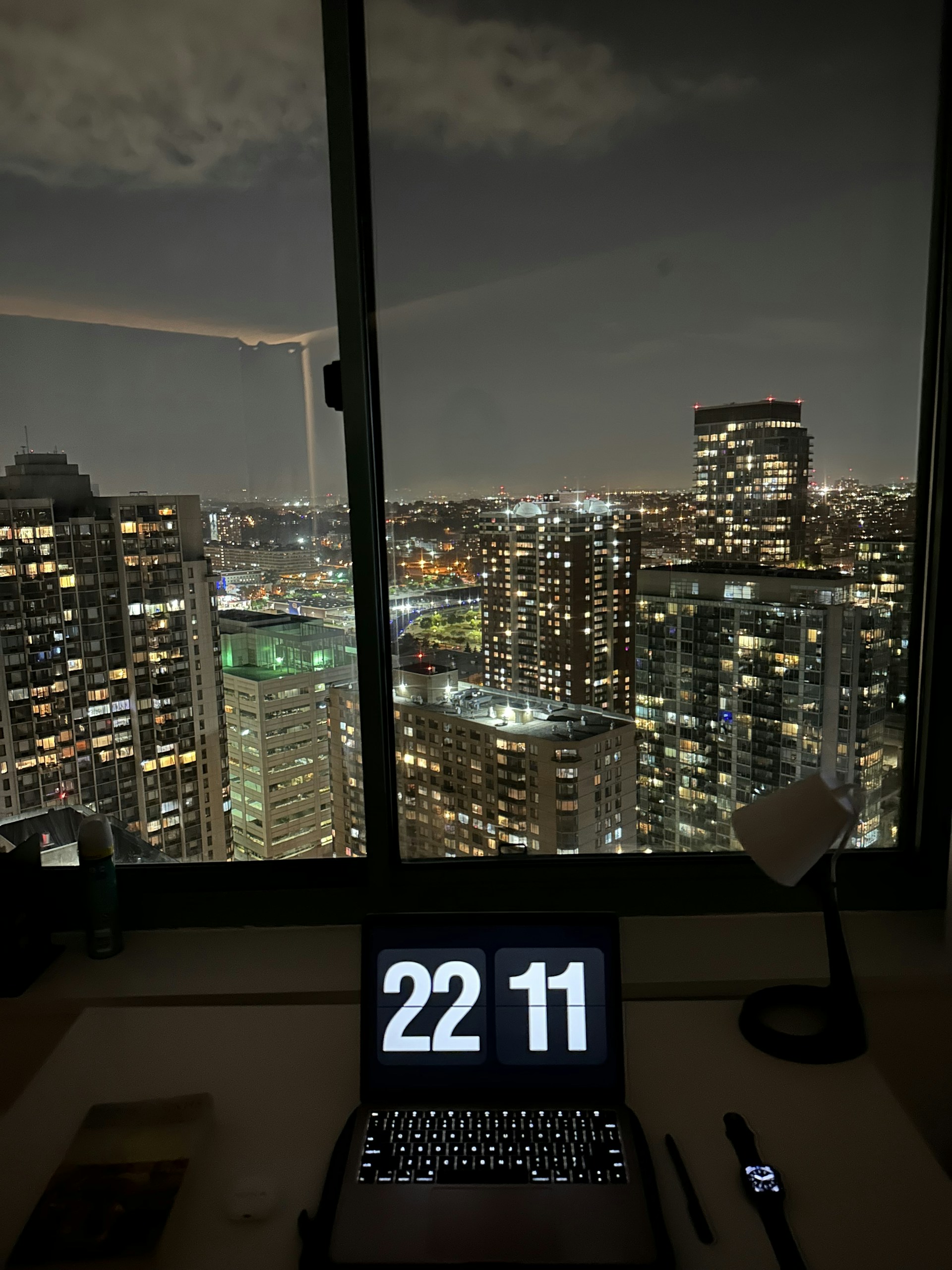 Close-up of hands typing on a laptop keyboard, with city lights beginning to twinkle outside the skyscraper window behind.