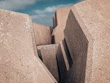Large geometric concrete blocks with rough surfaces form a structure resembling a barrier or breakwater. The edges are angular and the texture is rough with small white spots scattered across the surface. The sky in the background is partly cloudy.