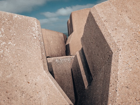 Large geometric concrete blocks with rough surfaces form a structure resembling a barrier or breakwater. The edges are angular and the texture is rough with small white spots scattered across the surface. The sky in the background is partly cloudy.