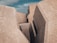 Large geometric concrete blocks with rough surfaces form a structure resembling a barrier or breakwater. The edges are angular and the texture is rough with small white spots scattered across the surface. The sky in the background is partly cloudy.
