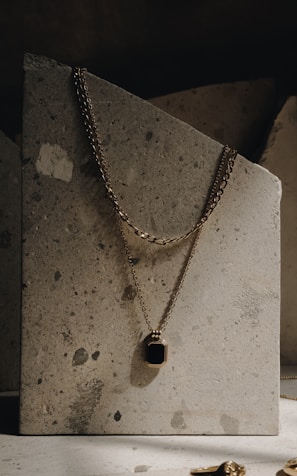 An elegant gold necklace with a rectangular black pendant is draped over a textured stone slab. The necklace's chain is visible, hanging gracefully against the rough, speckled surface. The lighting is soft and creates a subtle shadow, enhancing the luxurious feel.