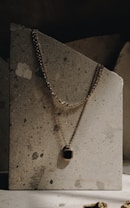 An elegant gold necklace with a rectangular black pendant is draped over a textured stone slab. The necklace's chain is visible, hanging gracefully against the rough, speckled surface. The lighting is soft and creates a subtle shadow, enhancing the luxurious feel.