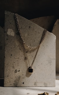 An elegant gold necklace with a rectangular black pendant is draped over a textured stone slab. The necklace's chain is visible, hanging gracefully against the rough, speckled surface. The lighting is soft and creates a subtle shadow, enhancing the luxurious feel.