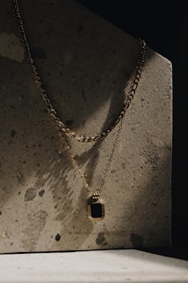 A gold chain necklace with a rectangular pendant featuring a black stone is draped against a textured concrete surface. The lighting is moody with shadows creating a dramatic effect on the surface.