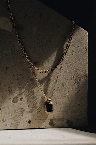 A gold chain necklace with a rectangular pendant featuring a black stone is draped against a textured concrete surface. The lighting is moody with shadows creating a dramatic effect on the surface.