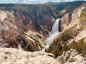 A breathtaking view of a waterfall cascading down a steep cliff surrounded by dense forest. The waterfall creates a mist at its base, and the cliffs exhibit layers of colorful sediment. The background consists of a lush forest under a partially cloudy sky, offering a sense of tranquility and grandeur.