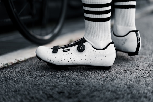 A close-up of a cyclist's feet wearing white cycling shoes with black accents and standing on a road with textured pavement. The cyclist is also wearing white socks with black stripes. A bicycle wheel is partially visible in the background.
