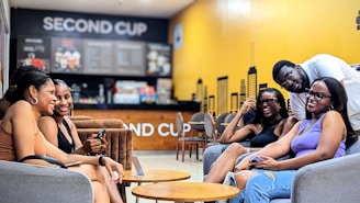 A group of five people are sitting in a cozy coffee shop, engaging in conversation and laughter. The background features a counter with a menu board displaying various drink options. The coffee shop has a bright yellow wall with black accents and the words 'SECOND CUP' prominently displayed.