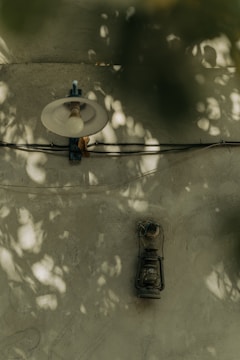 A lamp with a large bulb is affixed to a rough, textured wall. The surface is illuminated by dappled sunlight, creating scattered patterns of light and shadow. Below the lamp, an old-fashioned hanging lantern is mounted. Both the lamp and the lantern are connected by a wire running along the wall.