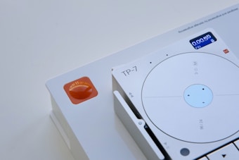A close-up of a modern electronic device featuring a circular dial with a small digital display in the top right, showing the time 0:00:55 and a day indicator. The device is predominantly white with minimalist design elements and the letters 'TP-7' are visible near the dial. An orange, semi-transparent button or indicator is positioned on the left side of the image.
