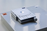 A sleek, modern electronic device with a circular interface and several buttons prominently displayed on a smooth surface. The device appears to have a minimalist design with a subtle light indicator and a textured knob on one side.