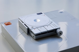 A sleek, modern electronic device with a circular interface and several buttons prominently displayed on a smooth surface. The device appears to have a minimalist design with a subtle light indicator and a textured knob on one side.
