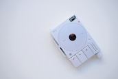 A minimalist electronic device with a white and gray color scheme, featuring a circular central component and several buttons and indicators. The display shows digital information, and there are various controls for functionality, including a large knob.