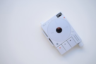 A minimalist electronic device with a white and gray color scheme, featuring a circular central component and several buttons and indicators. The display shows digital information, and there are various controls for functionality, including a large knob.