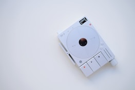 A minimalist electronic device with a white and gray color scheme, featuring a circular central component and several buttons and indicators. The display shows digital information, and there are various controls for functionality, including a large knob.
