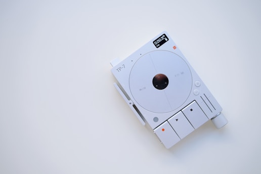 A minimalist electronic device with a white and gray color scheme, featuring a circular central component and several buttons and indicators. The display shows digital information, and there are various controls for functionality, including a large knob.