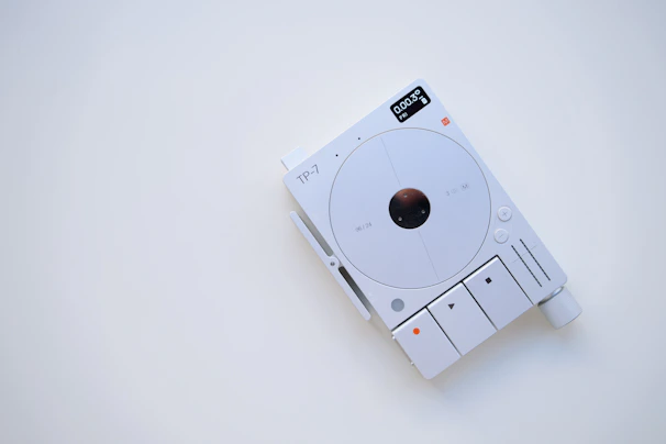 A minimalist electronic device with a white and gray color scheme, featuring a circular central component and several buttons and indicators. The display shows digital information, and there are various controls for functionality, including a large knob.