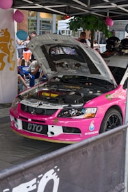 A bright pink rally car with its hood open, revealing the engine. It's parked under a canopy with people gathered around, some wearing helmets. Decorations include balloons and banners in the background, adding a festive atmosphere.