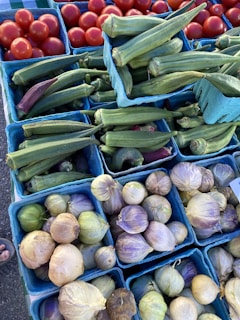 An assortment of fresh produce in small blue baskets, including red tomatoes, green okra, and husk-covered tomatillos. The arrangement suggests a market or grocery display with vibrant and natural colors.