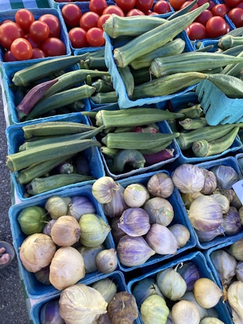 An assortment of fresh produce in small blue baskets, including red tomatoes, green okra, and husk-covered tomatillos. The arrangement suggests a market or grocery display with vibrant and natural colors.