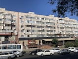 A multi-story residential building with a facade of uniform balconies and windows, featuring air conditioning units. The ground level includes commercial businesses with visible signage and a small restaurant terrace. Several cars are parked or driving on the road in front.