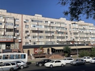A multi-story residential building with a facade of uniform balconies and windows, featuring air conditioning units. The ground level includes commercial businesses with visible signage and a small restaurant terrace. Several cars are parked or driving on the road in front.
