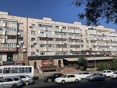 A multi-story residential building with a facade of uniform balconies and windows, featuring air conditioning units. The ground level includes commercial businesses with visible signage and a small restaurant terrace. Several cars are parked or driving on the road in front.