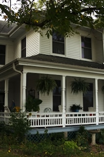 A two-story house with white siding and a gabled roof features a cozy front porch adorned with hanging ferns and potted plants. Wooden chairs and a small table are arranged neatly on the porch, surrounded by lush greenery.