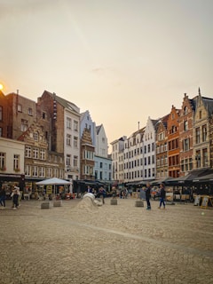 A picturesque European square with cobblestone pavement surrounded by historic buildings with unique gabled roofs. People are walking and gathering, while cafes with outdoor seating line the square, suggesting a lively atmosphere. The sky is clear with a warm light from the setting sun.