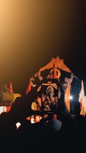 A smartphone held in the foreground captures an image of a brightly lit statue of a deity with an elephant head, possibly part of a religious procession. Various figures are visible surrounding the statue, which is adorned with orange decorations. The scene is set against a dark background, with warm light casting a golden hue.