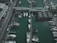 An aerial view of a marina with multiple boats and yachts docked alongside wooden piers. The area includes buildings with modern architecture and a bridge connecting different sections of the marina.
