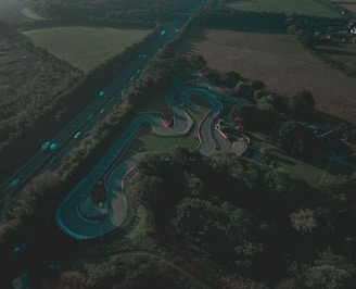An aerial view of a winding go-kart track surrounded by lush greenery and trees, with a highway to the side that has several vehicles traveling on it. The scene appears to be set during the day with long shadows cast by the trees, indicating early morning or late afternoon.